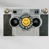 Shot on paper shoot text lens cover with yellow smiley face graphic with grip groves on a paper shoot camera 